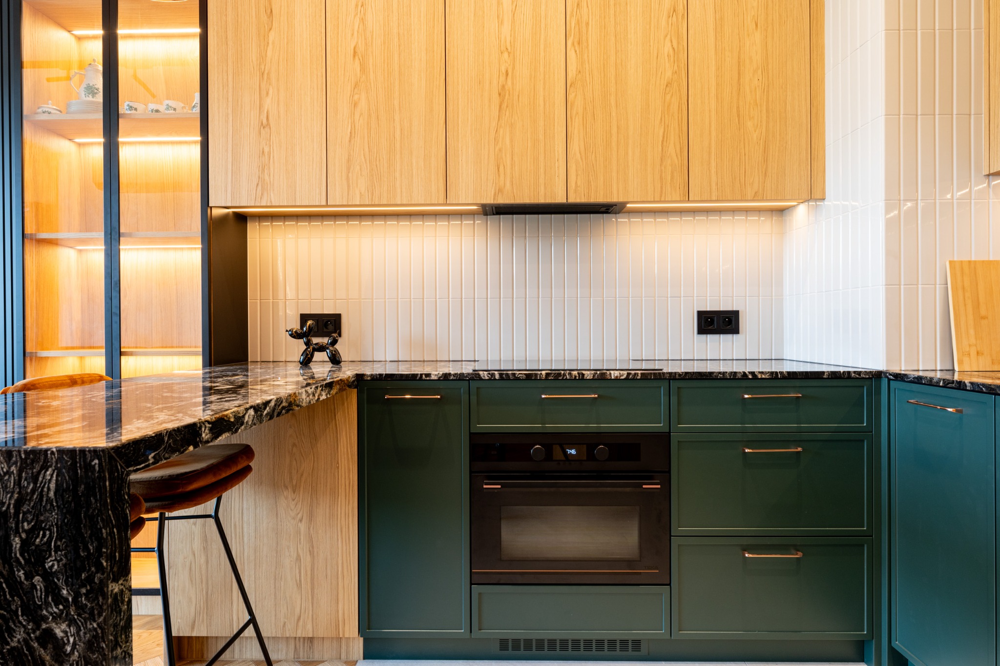Nowoczesna kuchnia z zielonymi szafkami, drewnianymi frontami i blatem z czarnego marmuru. Oświetlenie LED podkreśla detale. Na blacie czarny piesek.