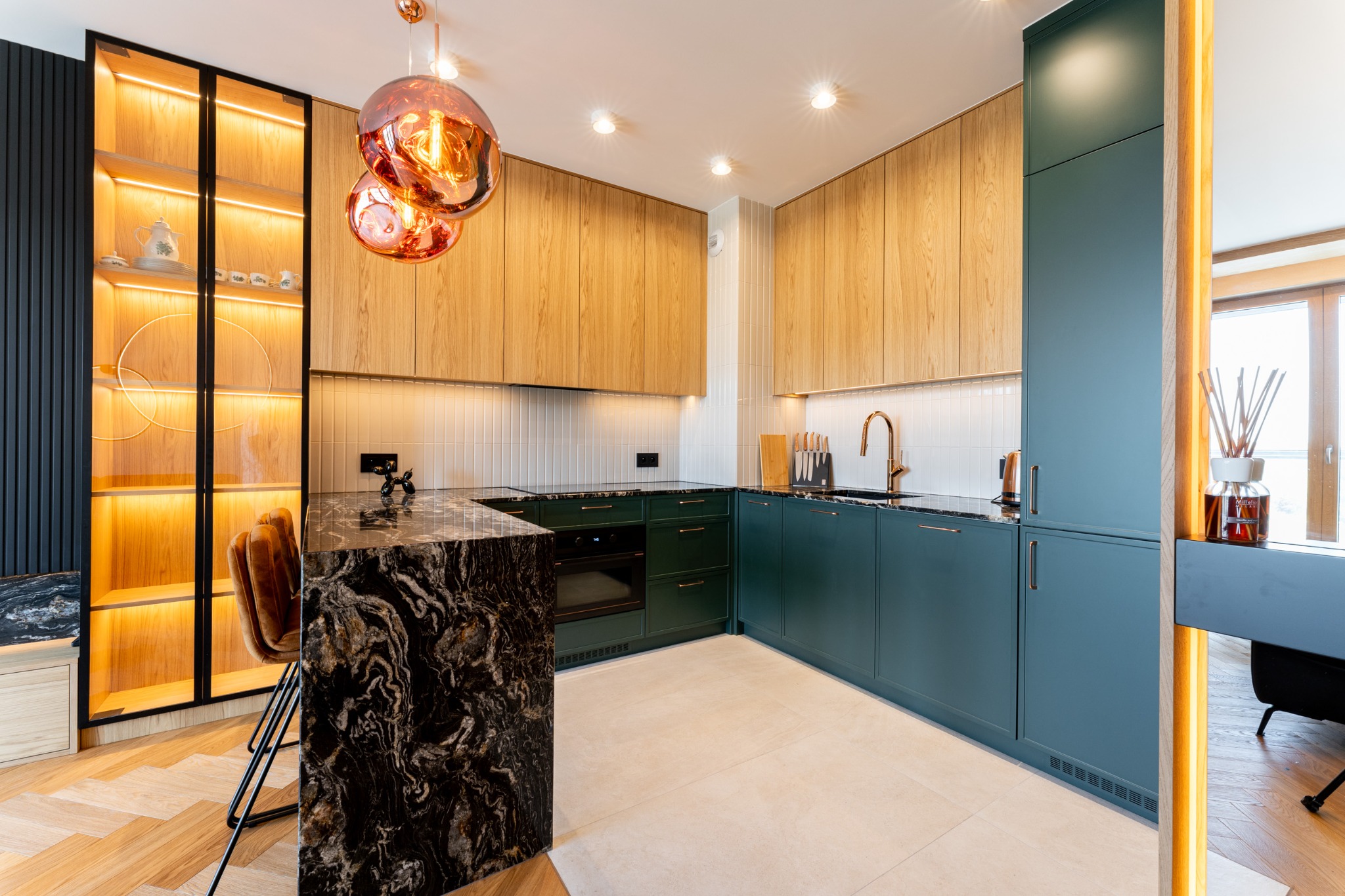 Nowoczesna kuchnia z zielonymi szafkami, drewnianymi frontami i blatem z czarnego marmuru. Oświetlona witryna z dekoracyjną porcelaną i lampy w stylu industrialnym.