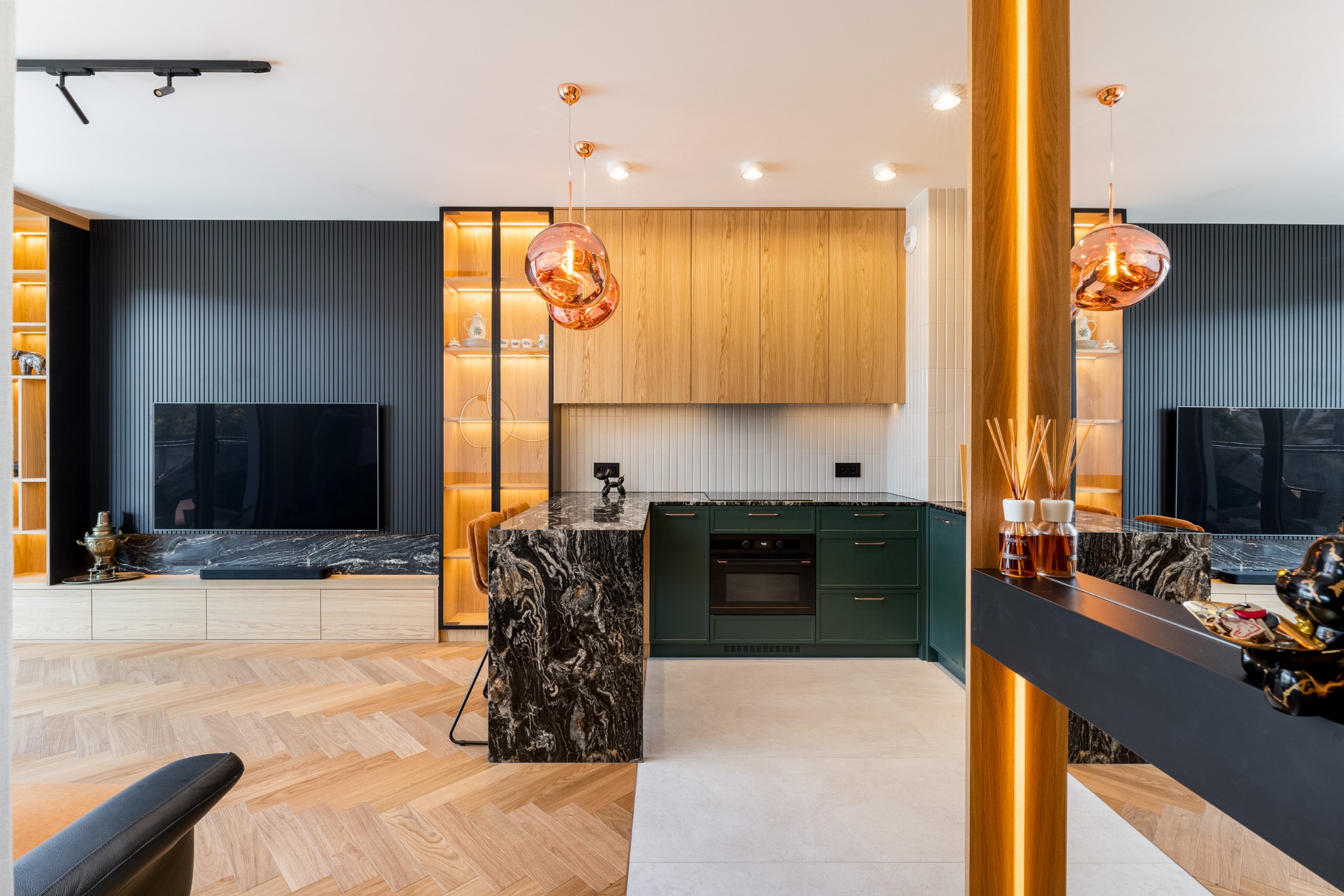 Nowoczesna kuchnia z zielonymi szafkami, blatem z czarnego marmuru i oświetleniem LED. Widoczne elementy dekoracyjne i drewniane wykończenia. Uwagę przykuwają lampy wiszące.