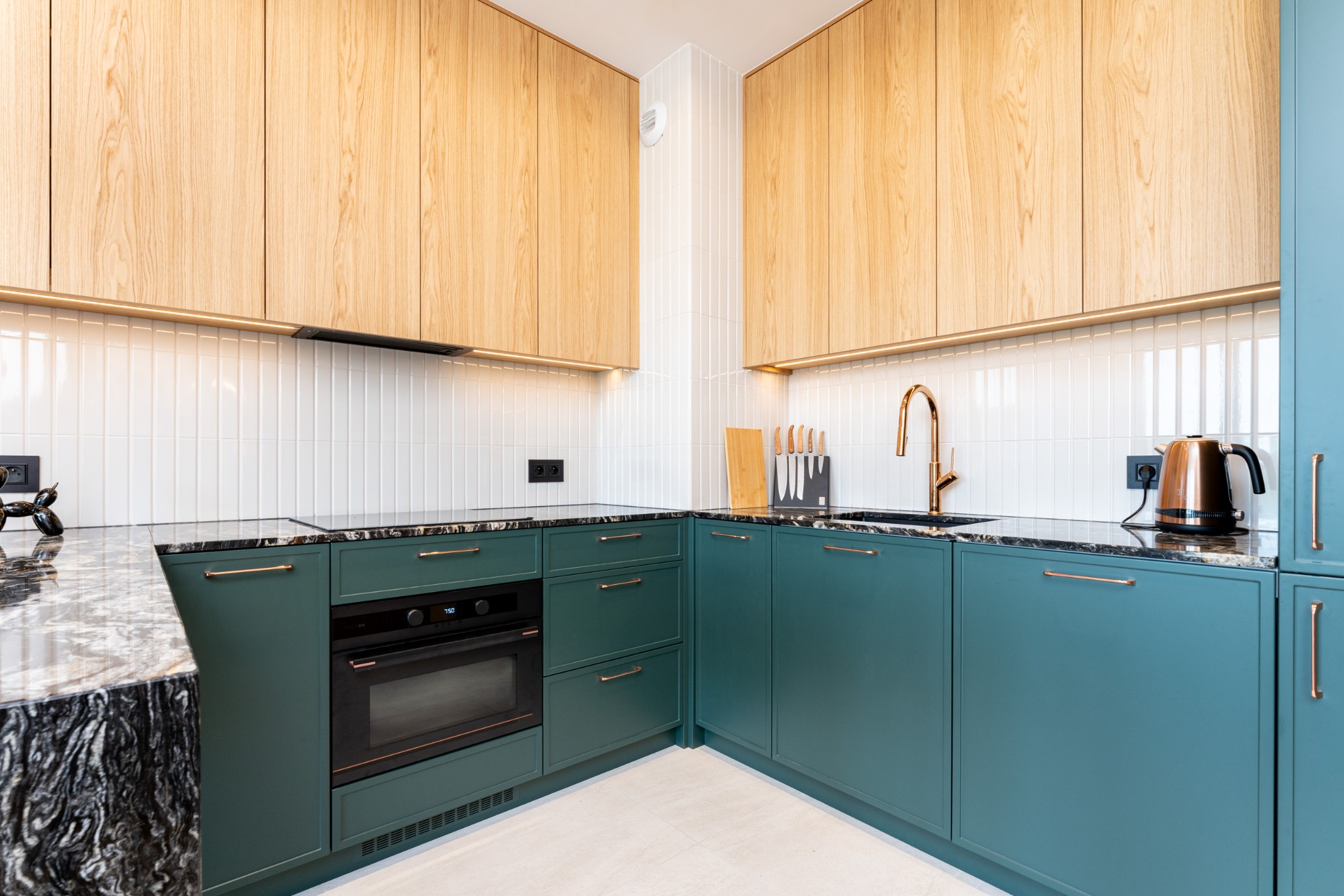 Nowoczesna kuchnia z zielonymi szafkami dolnymi i drewnianymi górnymi, czarnym blatem i złotymi detalami, oświetlona listwami LED pod szafkami.