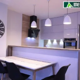 Nowoczesna kuchnia z białymi frontami i drewnianymi akcentami, oświetlona trzema wiszącymi lampami, z widocznym dużym zegarem ściennym i stołem jadalnym z czarnymi krzesłami.