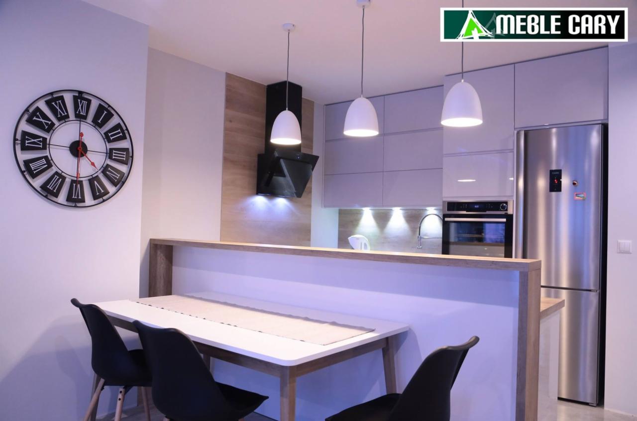 Nowoczesna kuchnia z białymi frontami i drewnianymi akcentami, oświetlona trzema wiszącymi lampami, z widocznym dużym zegarem ściennym i stołem jadalnym z czarnymi krzesłami.