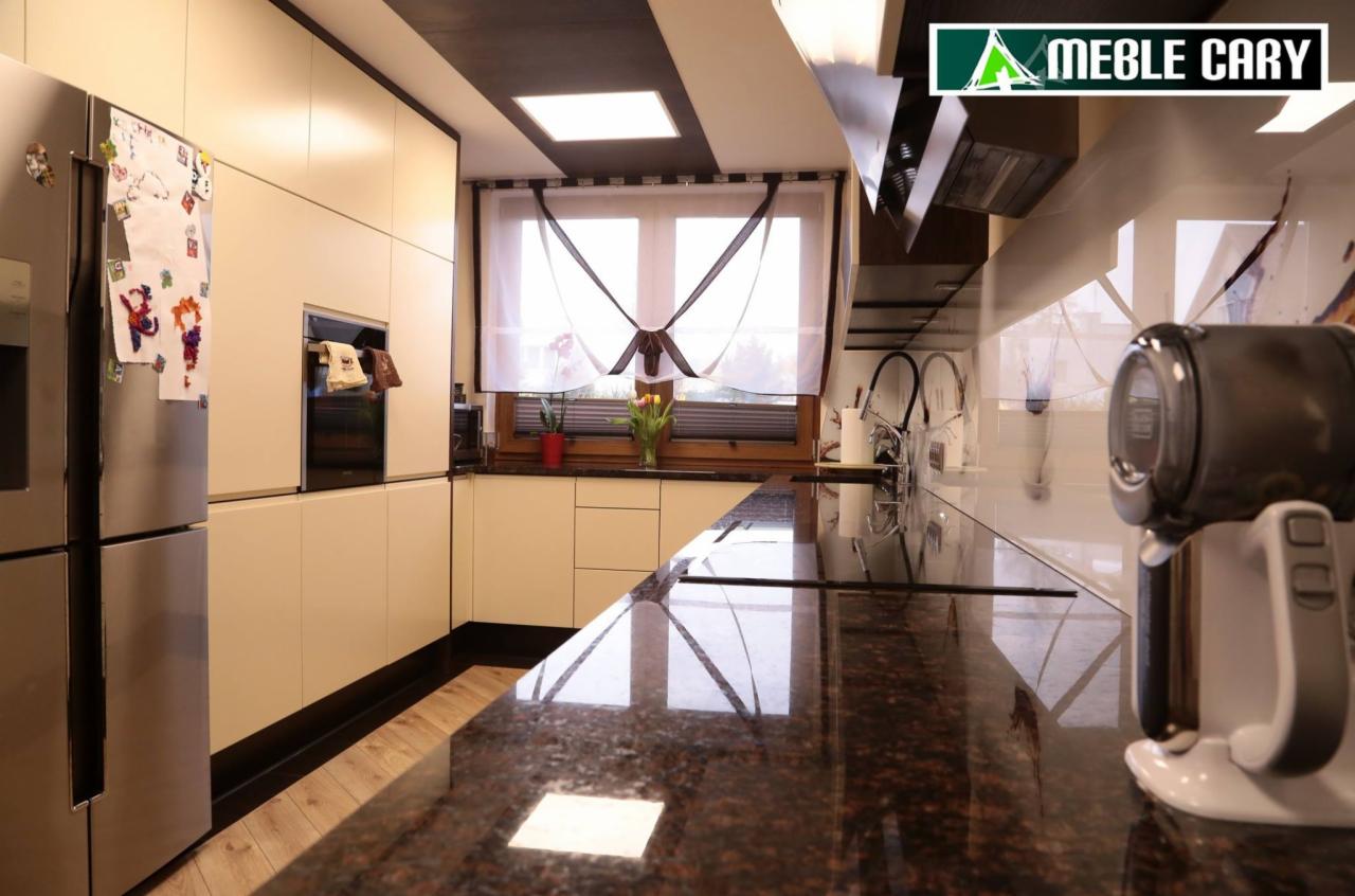 Nowoczesna kuchnia w jasnych barwach z ciemnym blatem, widoczny fragment lodówki z magnesami, okno z dekoracyjną zasłoną i sprzęt AGD na blacie.