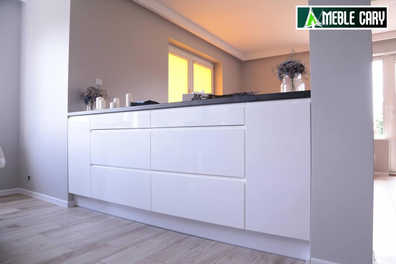 Białe, minimalistyczne szafki kuchenne z szufladami i czarnym blatem, dekorowane bukietem w szklanym wazonie, widok z niskiej perspektywy, logo firmy w prawym górnym rogu.