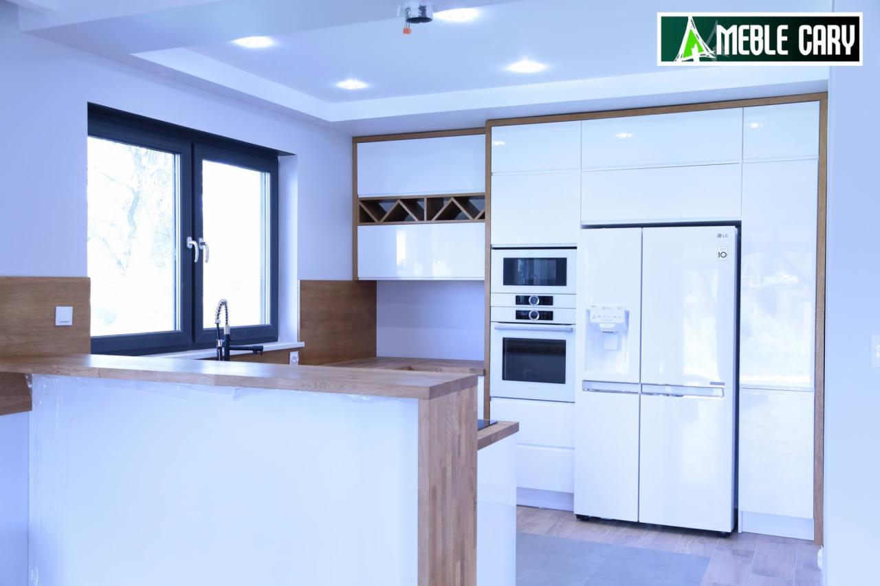 Nowoczesna, jasna kuchnia z białymi, lakierowanymi frontami, drewnianymi akcentami i wyspą; widoczny narożny barek, zabudowa AGD z lodówką side-by-side i oknem z czarną ramą.