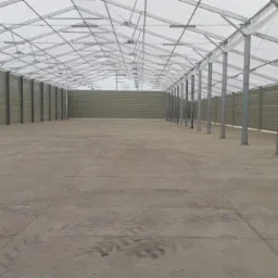 Wnętrze dużej hali z betonową posadzką, stalowymi słupami i ścianami z blachy trapezowej pod białym, półprzezroczystym dachem.