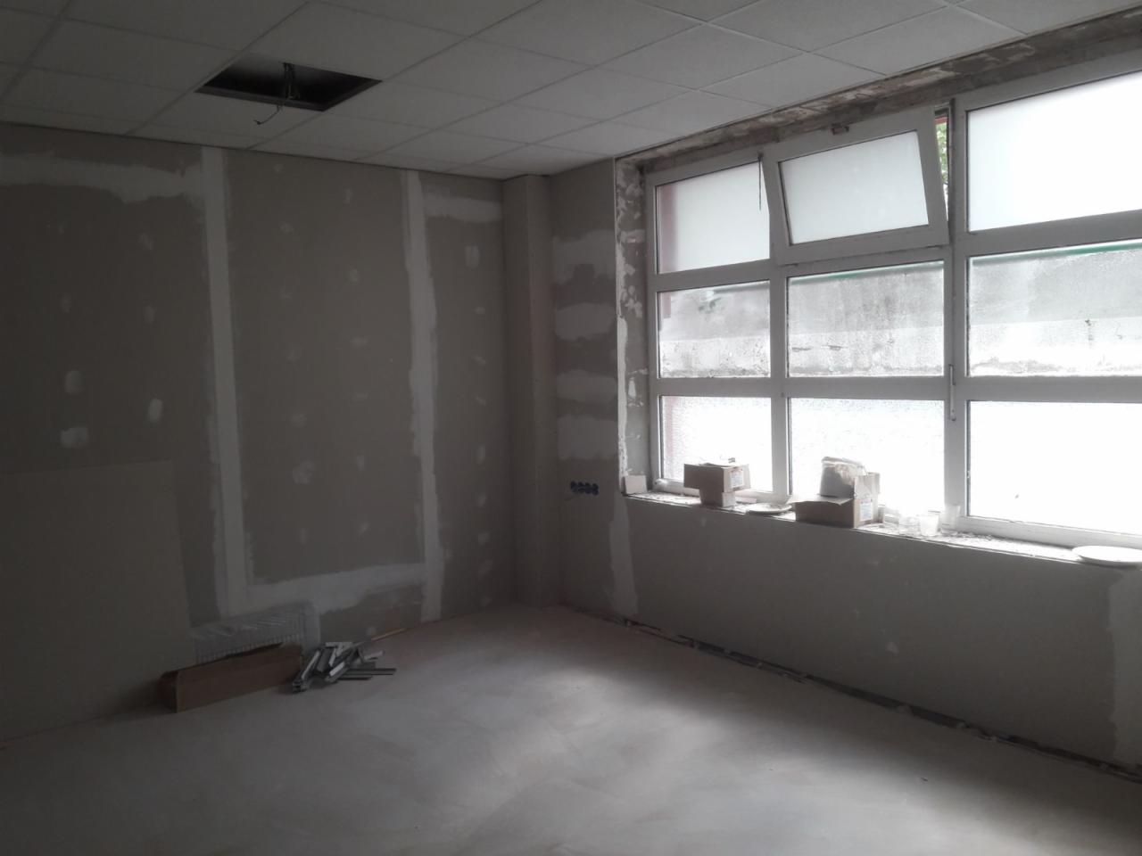 Wnętrze w trakcie remontu: ściany z płyt kartonowo-gipsowych przed szpachlowaniem, surowa wylewka podłogowa, okno z częściowo zamglonymi szybami, widoczne narzędzia i materiały budowlane na podłodze.