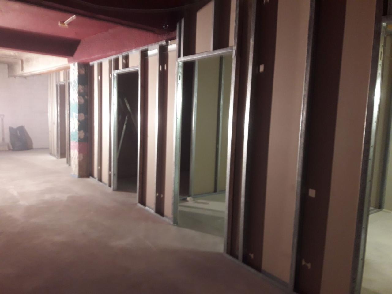 Montaż ścian działowych z płyt gipsowo-kartonowych w surowym stanie, widoczne metalowe profile i częściowo położona płyta, surowe wykończenie podłogi i stropu, widoczne instalacje pod sufitem.