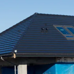 Nowy, ciemny dach z dwoma oknami połaciowymi na tle błękitnego nieba, widoczne rynny i fragment budynku w trakcie budowy, zabezpieczony niebieską folią.