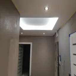 Korytarz w trakcie prac wykończeniowych, widoczne ściany pokryte gładzią, sufit z podwieszanym oświetleniem LED, zabezpieczone taśmą malarską ościeżnice drzwi.