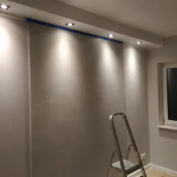 Świeżo pomalowane szare ściany z zamontowanymi punktowymi lampami sufitowymi, zabezpieczone niebieską taśmą malarską, widoczna drabina aluminiowa i okno.