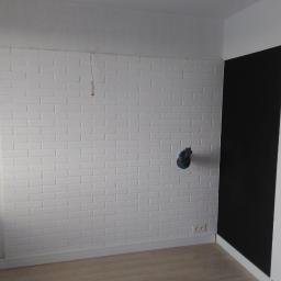 Tombud - Ściana z białej cegły dekoracyjnej w trakcie wykańczania, widoczne wystające kable elektryczne i otwór na gniazdko, obok czarny fragment ściany, jasna podłoga.