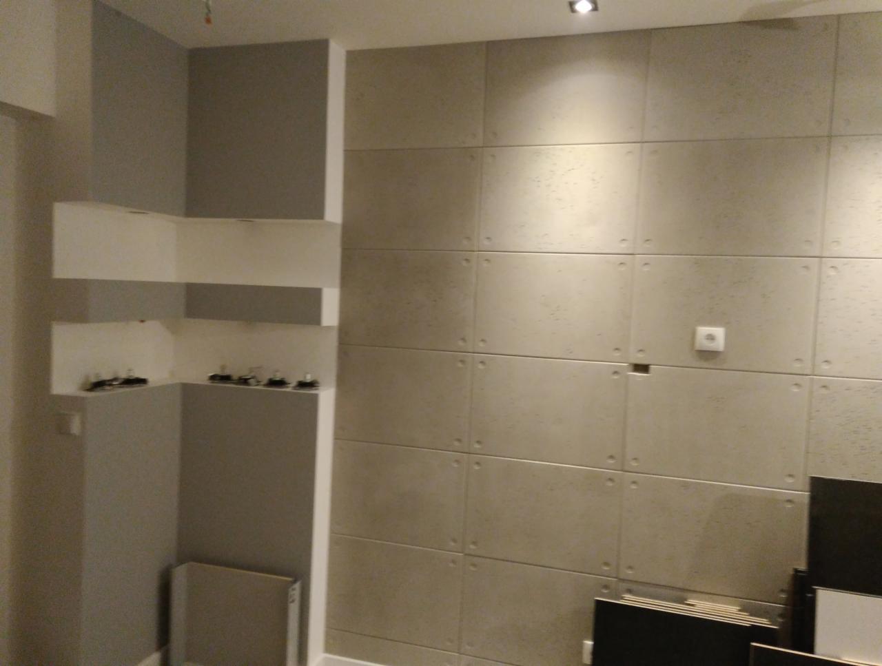 Ściana wykończona panelami imitującymi beton z widocznym gniazdkiem elektrycznym oraz półkami w narożniku, w trakcie prac wykończeniowych.