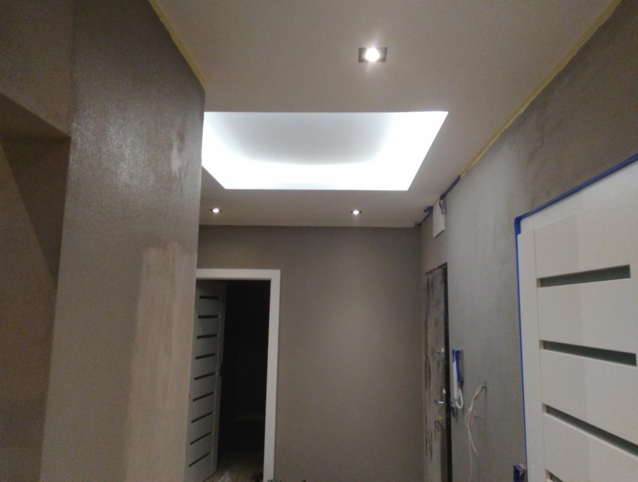 Korytarz w trakcie prac wykończeniowych, widoczne ściany pokryte gładzią, sufit z podwieszanym oświetleniem LED, zabezpieczone taśmą malarską ościeżnice drzwi.