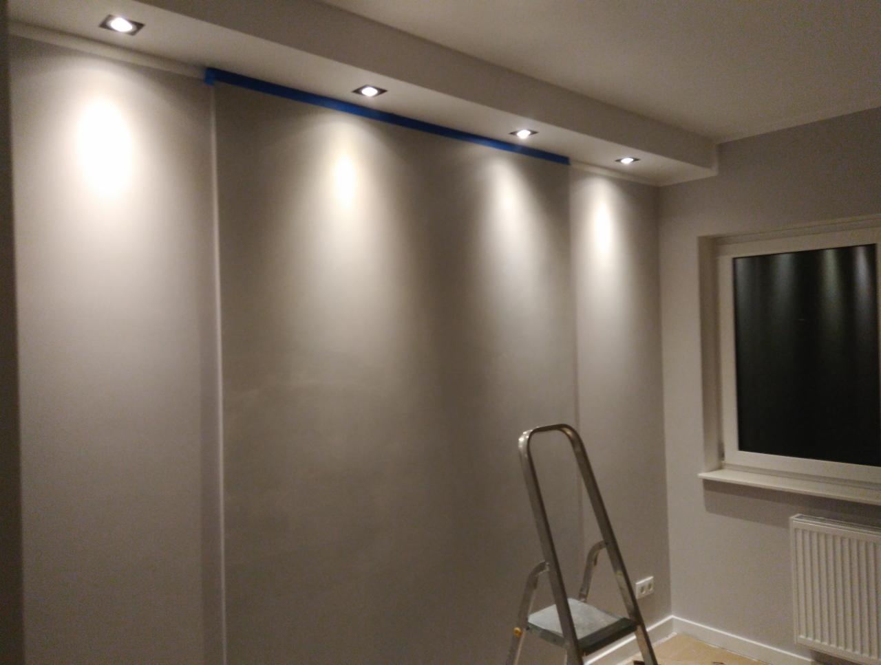 Świeżo pomalowane szare ściany z zamontowanymi punktowymi lampami sufitowymi, zabezpieczone niebieską taśmą malarską, widoczna drabina aluminiowa i okno.