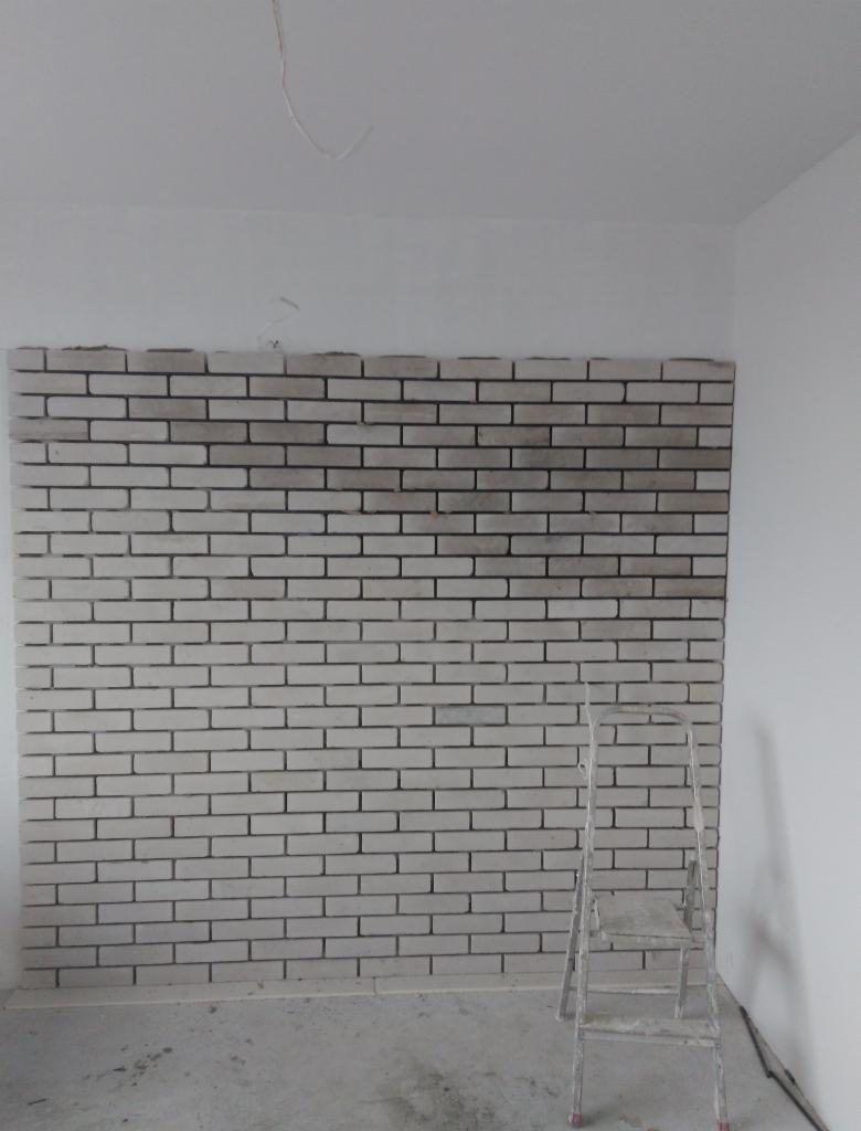 Ściana wyłożona częściowo białymi cegłami dekoracyjnymi z ciemną fugą, widoczna drabina aluminiowa, surowy stan wykończenia podłogi i ścian.