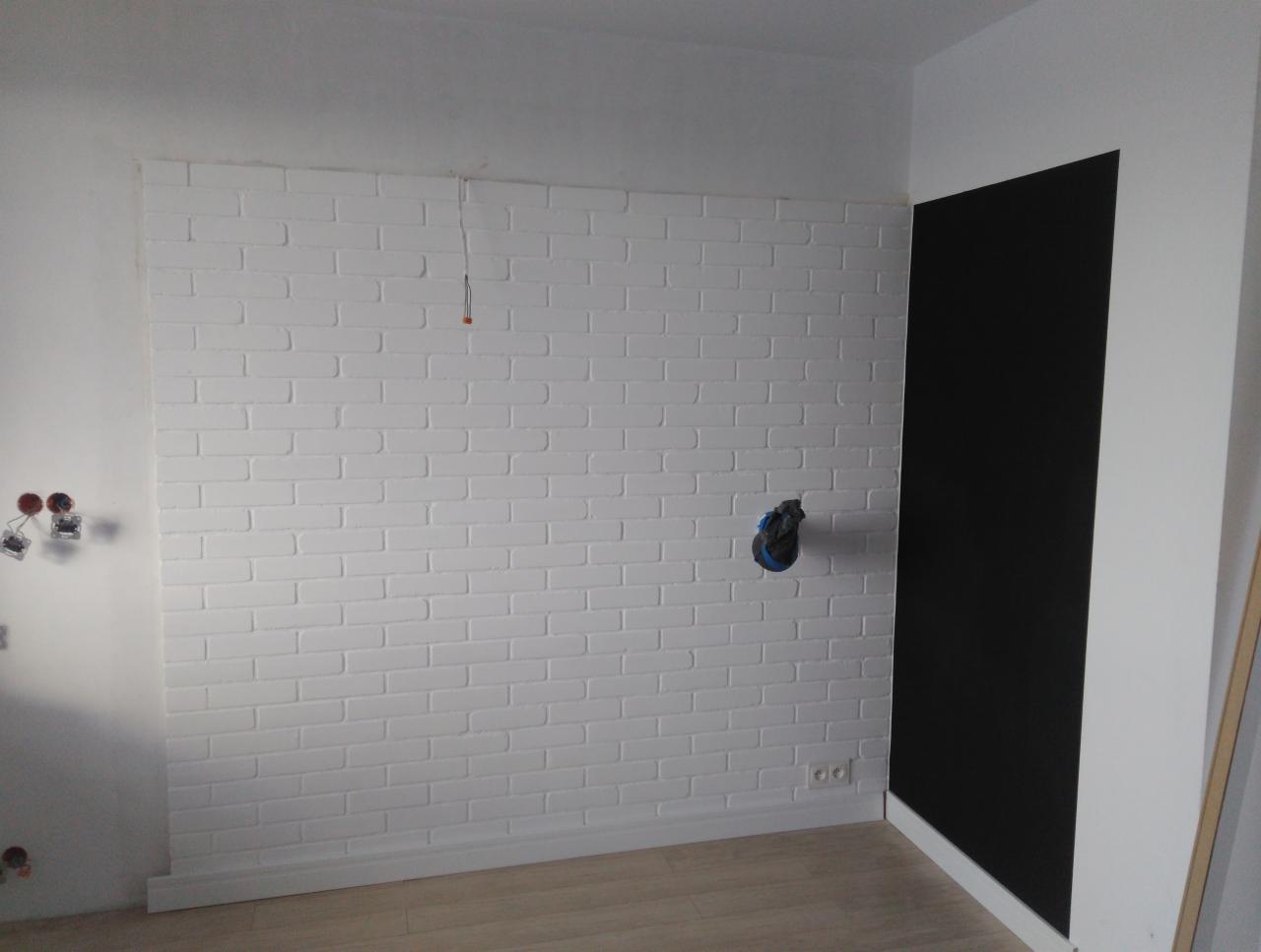 Ściana z białej cegły dekoracyjnej w trakcie wykańczania, widoczne wystające kable elektryczne i otwór na gniazdko, obok czarny fragment ściany, jasna podłoga.