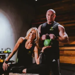 Uśmiechnięta blondynka siedzi na skrzyni w czarnym stroju sportowym, obok stoi muskularny mężczyzna trzymający zielony kettlebell 12 kg, oboje w koszulkach z logo 'Trener Paweł' na tle wyposażonej...