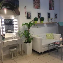 Wnętrze gabinetu kosmetycznego: toaletka z oświetleniem, pędzle do wizażu, biała sofa z zieloną poduszką, stolik kawowy ze świecą i czasopismami, roślina doniczkowa, zdjęcia i trofea na ścianie.