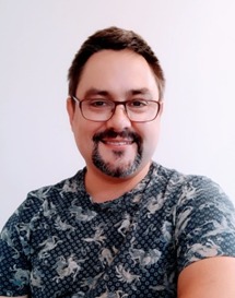 Portret mężczyzny w okularach i koszulce w abstrakcyjny wzór, z delikatnym uśmiechem, na jasnym tle.