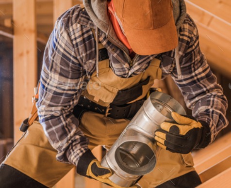 Instalator w roboczym ubraniu i rękawicach trzyma metalowy trójnik kanału wentylacyjnego na tle drewnianej konstrukcji dachu.