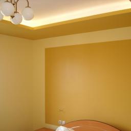 Ireneusz Osiecki - Żółte ściany w pokoju po malowaniu, widoczne gniazdka elektryczne i fragment łóżka, oświetlenie sufitowe z lampami kulistymi.