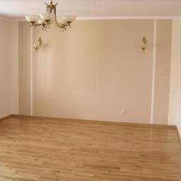 Ireneusz Osiecki - Pomalowane na beżowo ściany w pustym pokoju z parkietem, ozdobione klasycznym żyrandolem i kinkietami, biała listwa przypodłogowa i sufitowa.