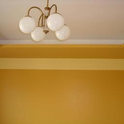 Ireneusz Osiecki - Ściana pomalowana na odcienie żółtego z dekoracyjnym obramowaniem i wiszącą lampą z białymi kulami.