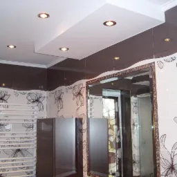 Wykończona łazienka z brązowo-białymi płytkami w kwiaty, podwieszanym sufitem z oświetleniem punktowym, grzejnikiem drabinkowym i lustrem w ozdobnej ramie.