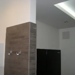 Fragment wnętrza łazienki z kolumną wyłożoną szarą płytką imitującą cegłę, na której zamontowano dwa chromowane wieszaki. Obok widoczna biała szafka instalacyjna i czarna szafka. Sufit...