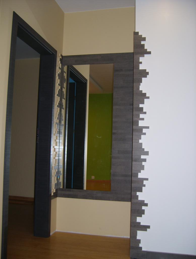 Fragment wnętrza: ciemna ościeżnica drzwiowa, lustro w ramie z szarej mozaiki przylegające do białej ściany z dekoracyjnym wykończeniem z mozaiki, widoczna zielona ściana w odbiciu lustra.