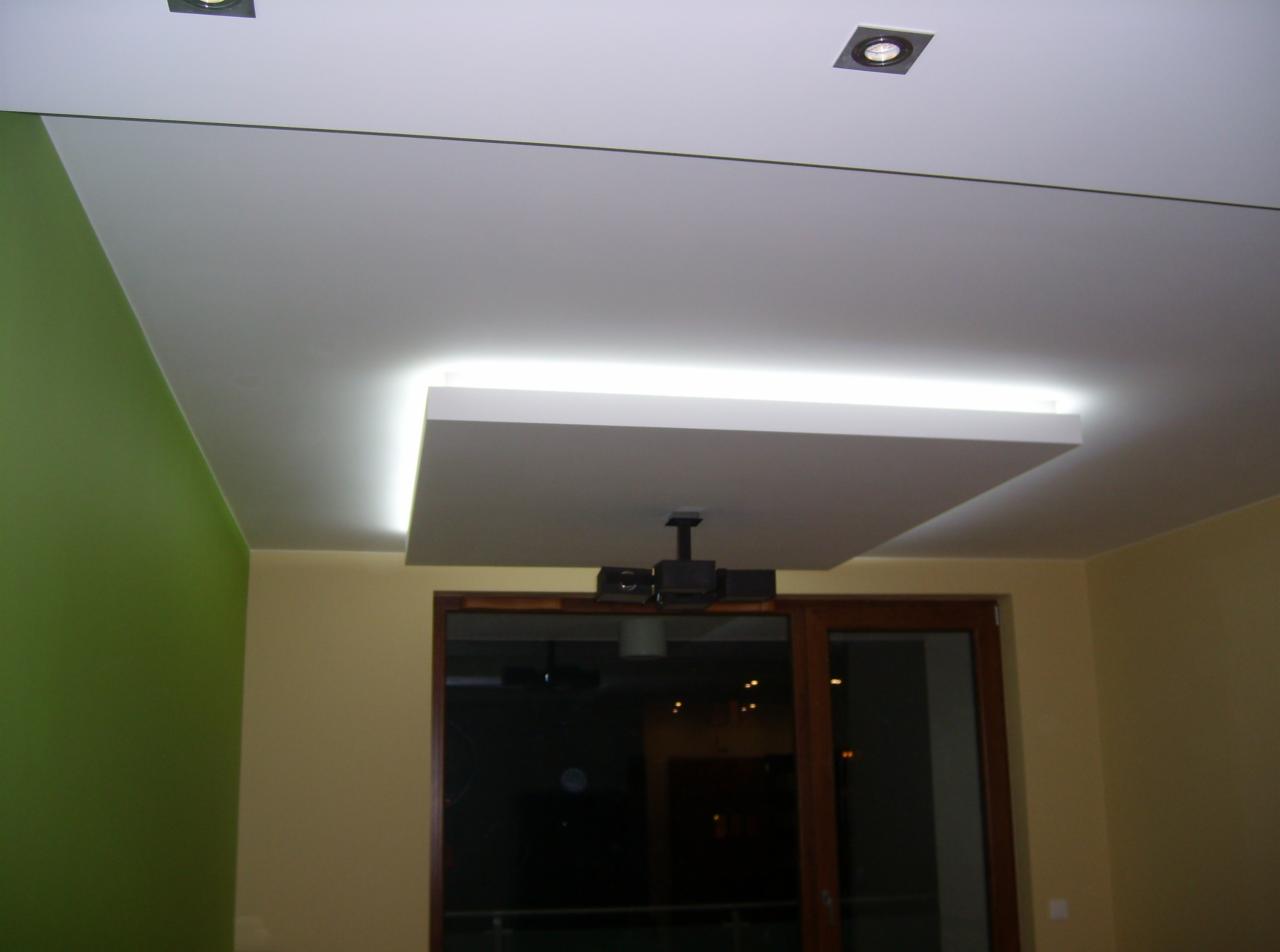 Nowoczesny sufit podwieszany z oświetleniem LED, widoczne również reflektory punktowe i fragment zielonej ściany, projektor zamontowany na suficie.