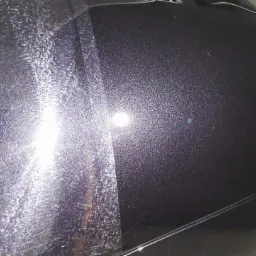 Część karoserii ciemnego samochodu przed i po polerowaniu, widoczne liczne zarysowania po lewej stronie i gładka powierzchnia po prawej.