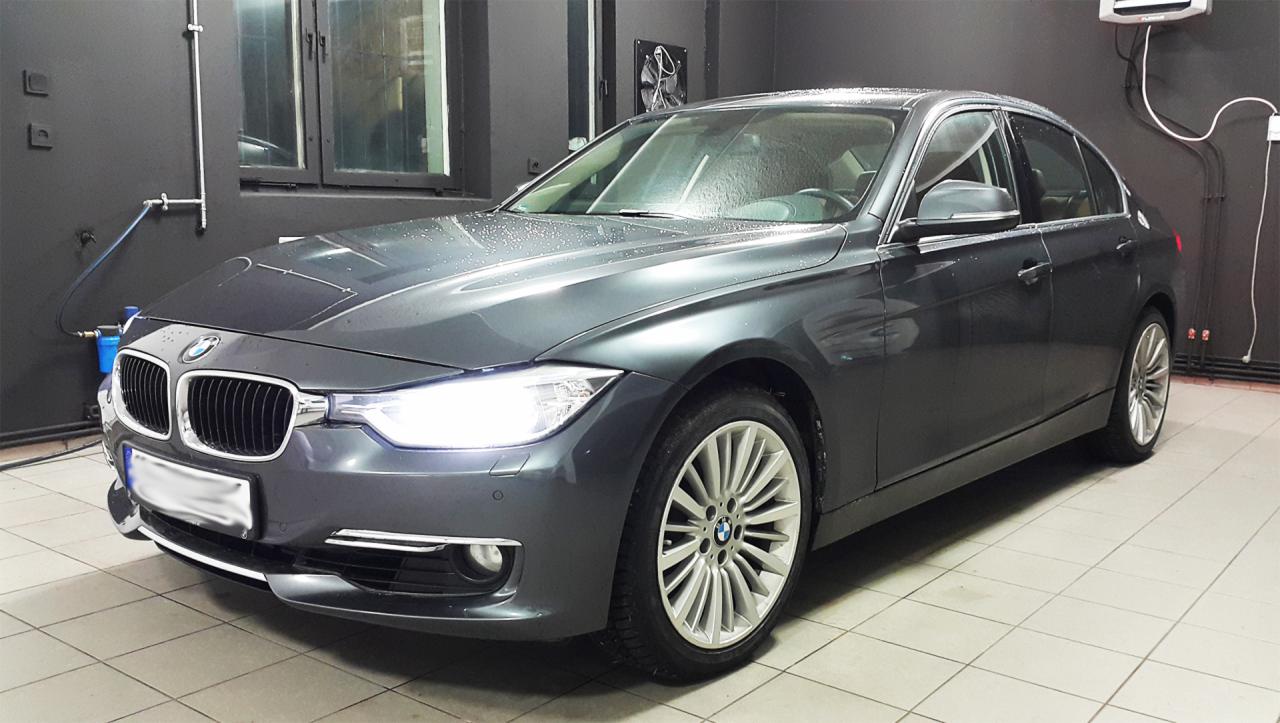 Szary samochód BMW serii 3 po umyciu, widoczne krople wody na karoserii, wnętrze myjni z szarymi ścianami i białymi płytkami podłogowymi.