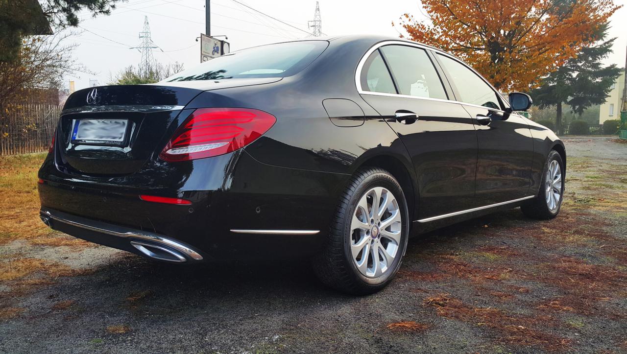 Błyszczący, czarny Mercedes E-Klasa po myciu, zaparkowany na żwirowej powierzchni, widok z tyłu, z widocznymi chromowanymi elementami wykończenia i aluminiowymi felgami.