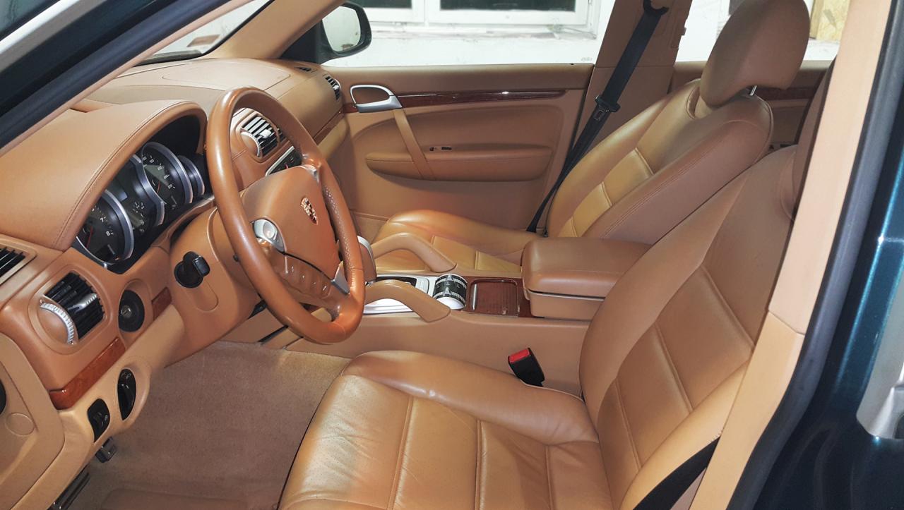 Wnętrze samochodu Porsche Cayenne z jasną skórzaną tapicerką, widok na deskę rozdzielczą, kierownicę z logo, fotele i konsolę środkową, drewniane wykończenia.
