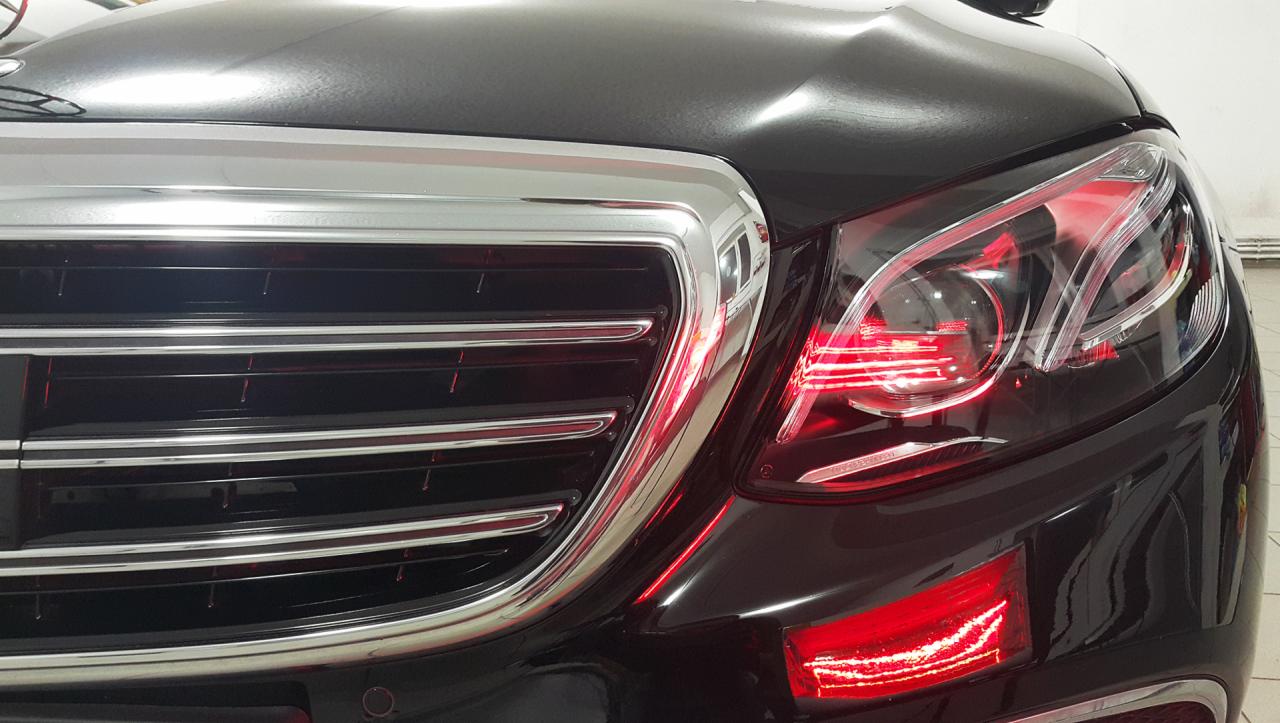 Błyszczący, czarny samochód osobowy marki Mercedes-Benz, widok z przodu na chromowaną atrapę chłodnicy i przedni reflektor z czerwonym światłem pozycyjnym.