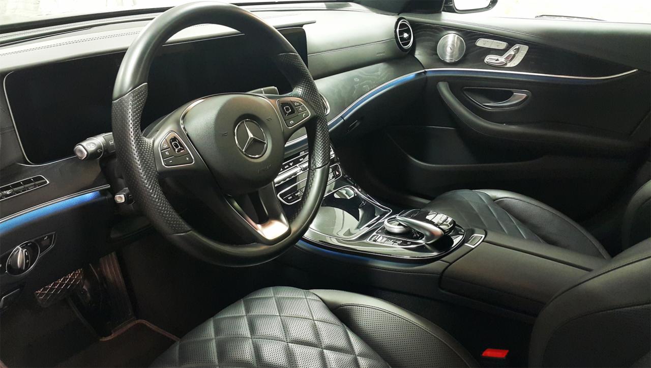 Wnętrze luksusowego samochodu Mercedes-Benz z czarną skórzaną tapicerką w romby, widoczne elementy deski rozdzielczej, kierownica z logo, srebrne detale i subtelne niebieskie podświetlenie ambientowe.