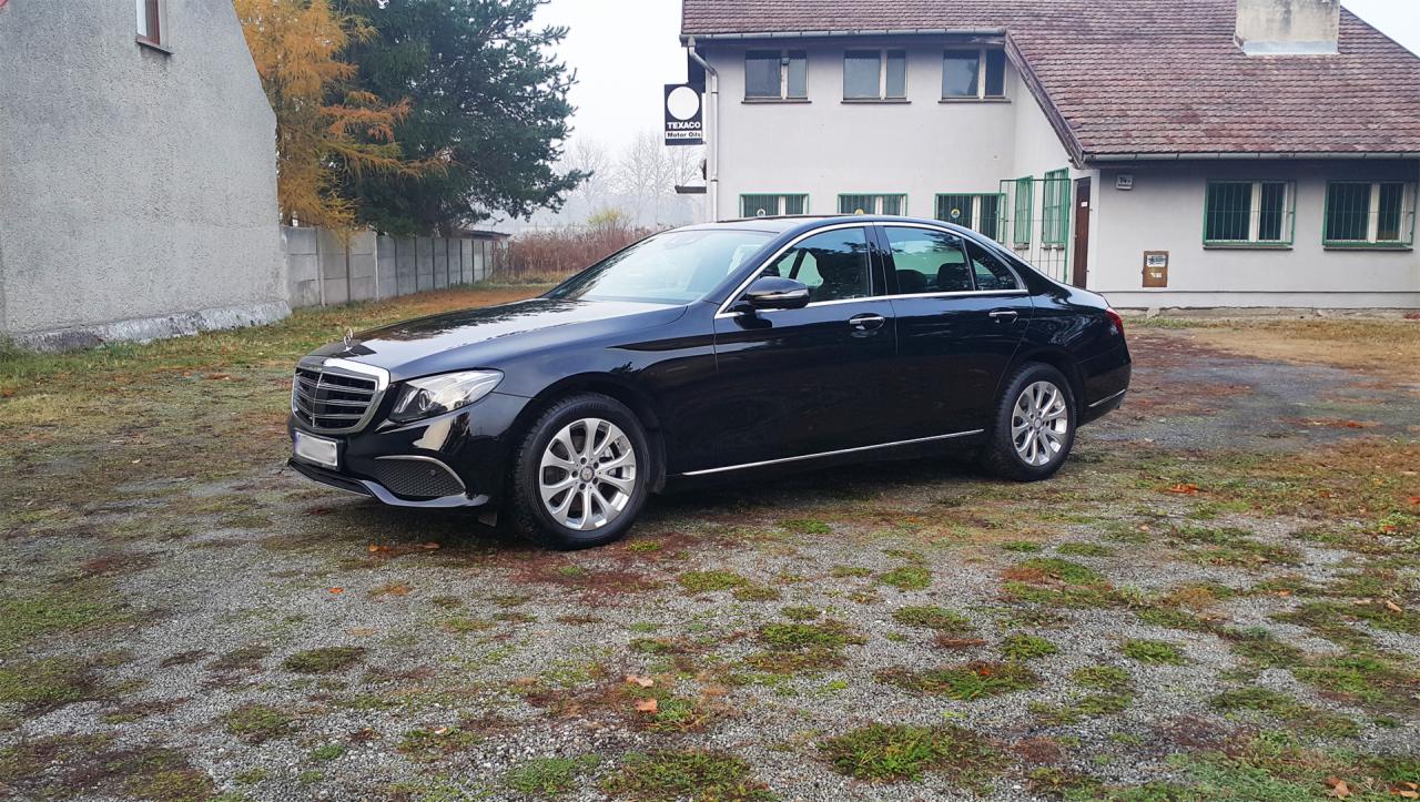 Czarny Mercedes-Benz klasy E zaparkowany na żwirowym podłożu przed budynkiem z logo Texaco w tle.