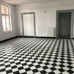 Wnętrze biura po remoncie z czarno-białą szachownicą na podłodze, widoczny grzejnik, okno z ceglaną ramą i sufit z rastrowymi oprawami oświetleniowymi.