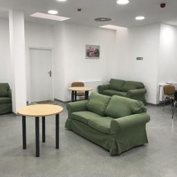 APTEBUD Rafał Szydlak - Jasne, przestronne wnętrze biurowe z zielonymi sofami, okrągłymi stolikami z ciemnymi nogami i obrazami kwiatów na ścianach, widoczne kratki wentylacyjne na suficie i znak ewakuacyjny.