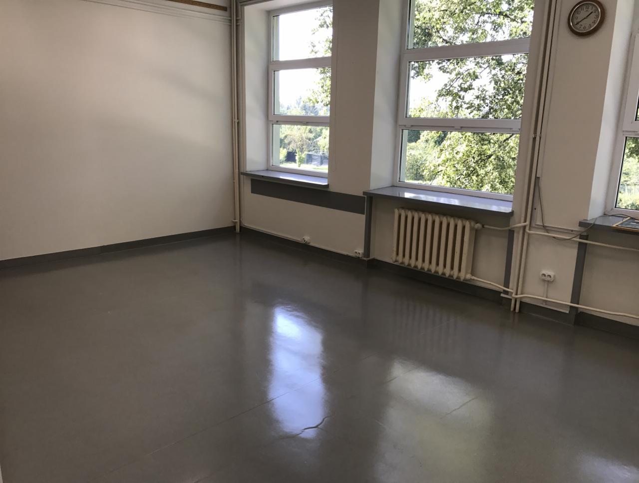Wnętrze pustego pomieszczenia z szarą podłogą, białymi ścianami, trzema oknami z widokiem na zieleń i starym grzejnikiem pod jednym z okien.