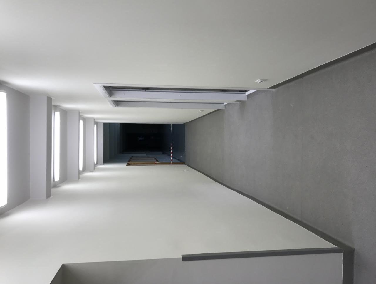 Wnętrze korytarza biurowego po remoncie, widok z góry, ściany pomalowane na szaro i biało, oświetlenie liniowe w kolumnach, sufit z otworem wentylacyjnym, zabezpieczona strefa prac na końcu korytarza.