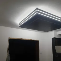 Sufit podwieszany z szarą płytą i trzema liniami oświetlenia LED, widok na fragment ściany z otworem drzwiowym w trakcie wykończenia.