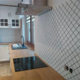 Nowoczesna kuchnia z białymi szafkami, drewnianym blatem, płytkami ściennymi w marokański wzór i czarną płytą indukcyjną; widoczny niedokończony montaż gniazdka elektrycznego.