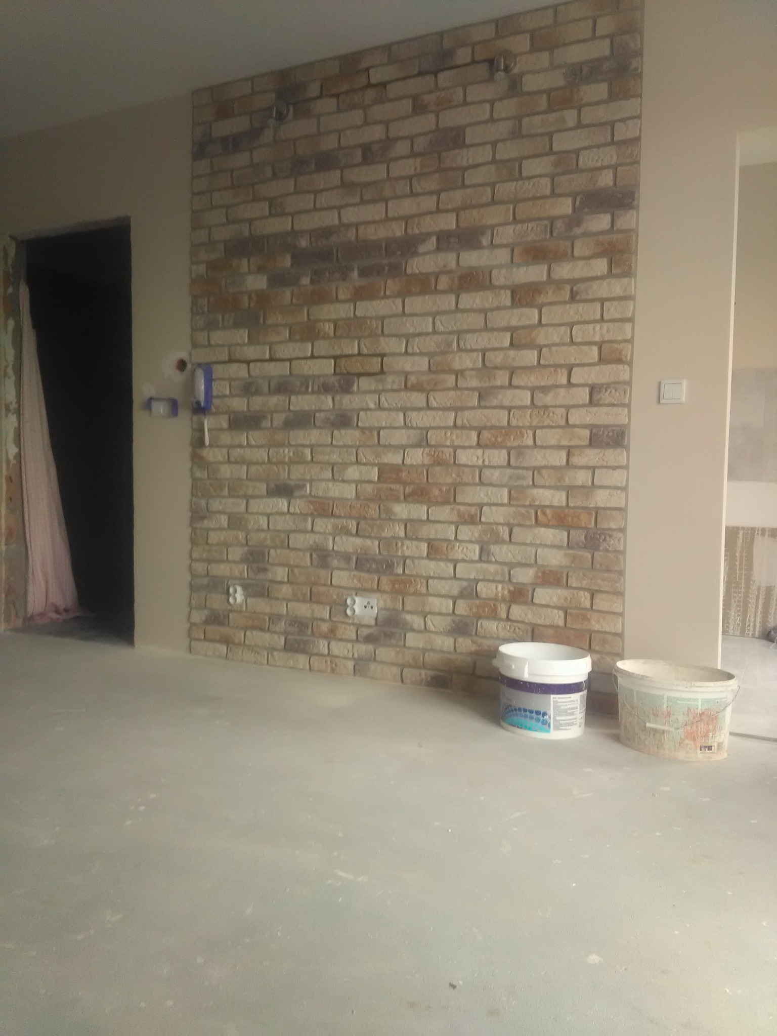Ściana wykończona cegłą dekoracyjną w odcieniach beżu i brązu, widoczne puszki elektryczne i dwa wiadra z farbą na surowej posadzce, sugerujące etap wykończeniowy.