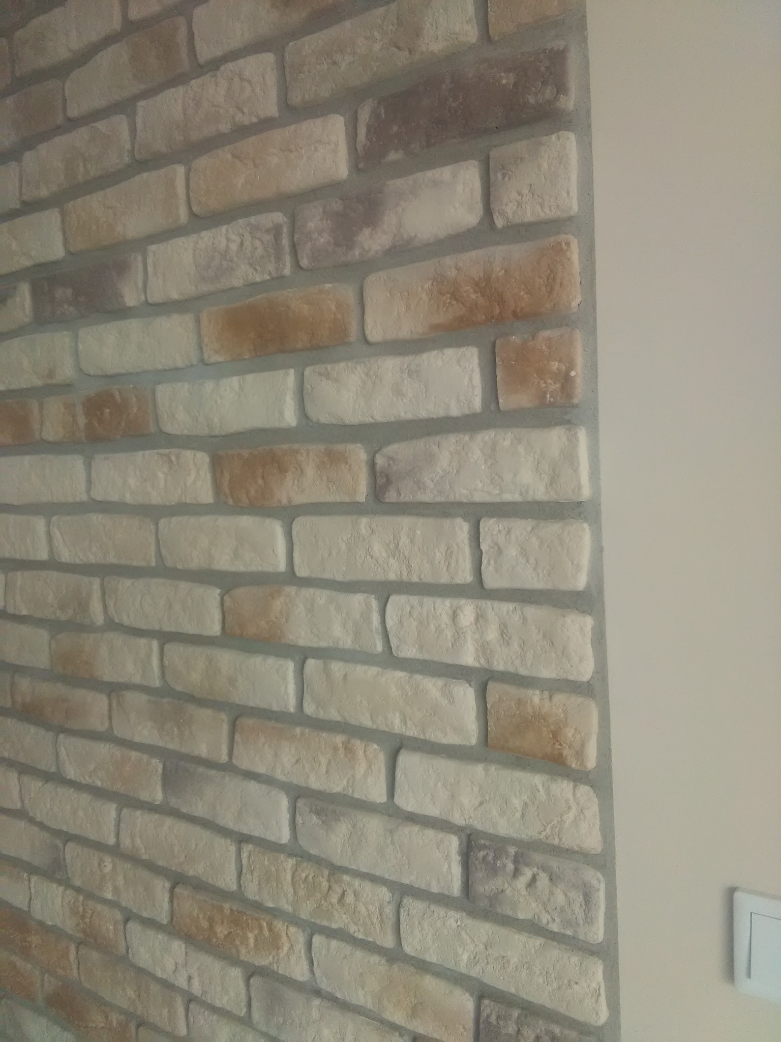 Ściana z dekoracyjnej cegły w odcieniach beżu i brązu, z widoczną fugą w kolorze szarym, przylegająca do gładkiej, pomalowanej ściany w jasnym odcieniu.