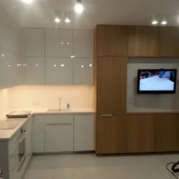 Nowoczesna kuchnia z białymi, błyszczącymi frontami i drewnianą zabudową z telewizorem, widok na umywalkę i płytę grzewczą, w tle drabina.