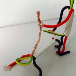 Kreatywna figurka człowieka uformowana z kolorowych przewod&oacute;w elektrycznych, z widocznymi miedzianymi żyłami, ustawiona na białym tle, prezentująca nietypowe zastosowanie materiał&oacute;w instalacyjnych.