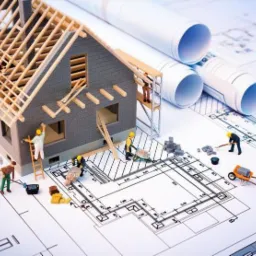 Makieta domu w trakcie budowy z miniaturowymi figurkami robotników, planami architektonicznymi w tle oraz zwiniętymi projektami, ukazująca proces wznoszenia konstrukcji.